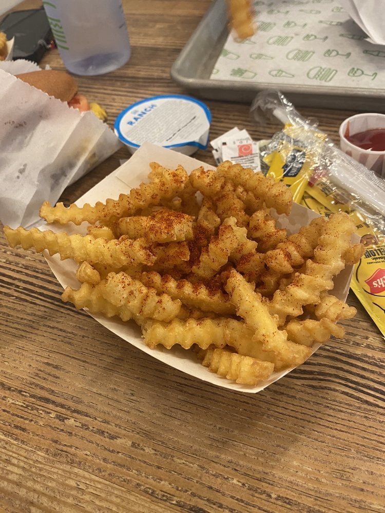 Crinkle-cut fries sprinkled with reddish seasoning served in a paper tray on a wooden table, with packets of condiments, a drink cup, and a burger partially visible in the background.