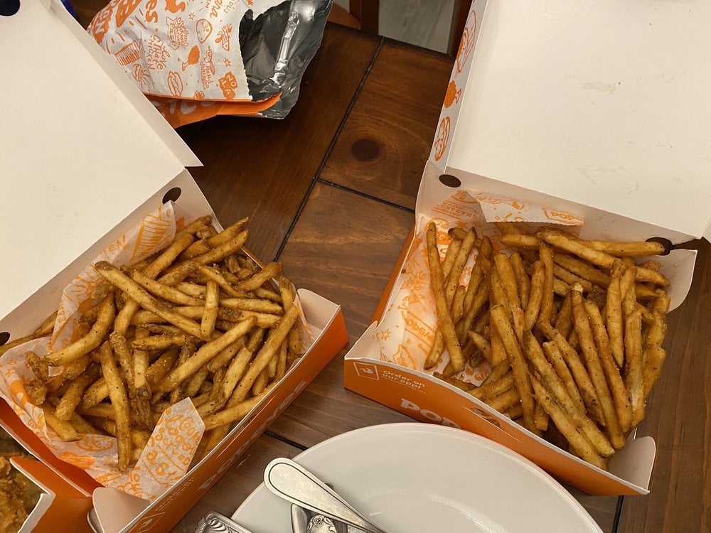 Two open boxes of Popeyes seasoned fries sit on a wooden table next to a plate with utensils. The fries are golden-brown and crispy, and the boxes are lined with branded Popeyes paper.