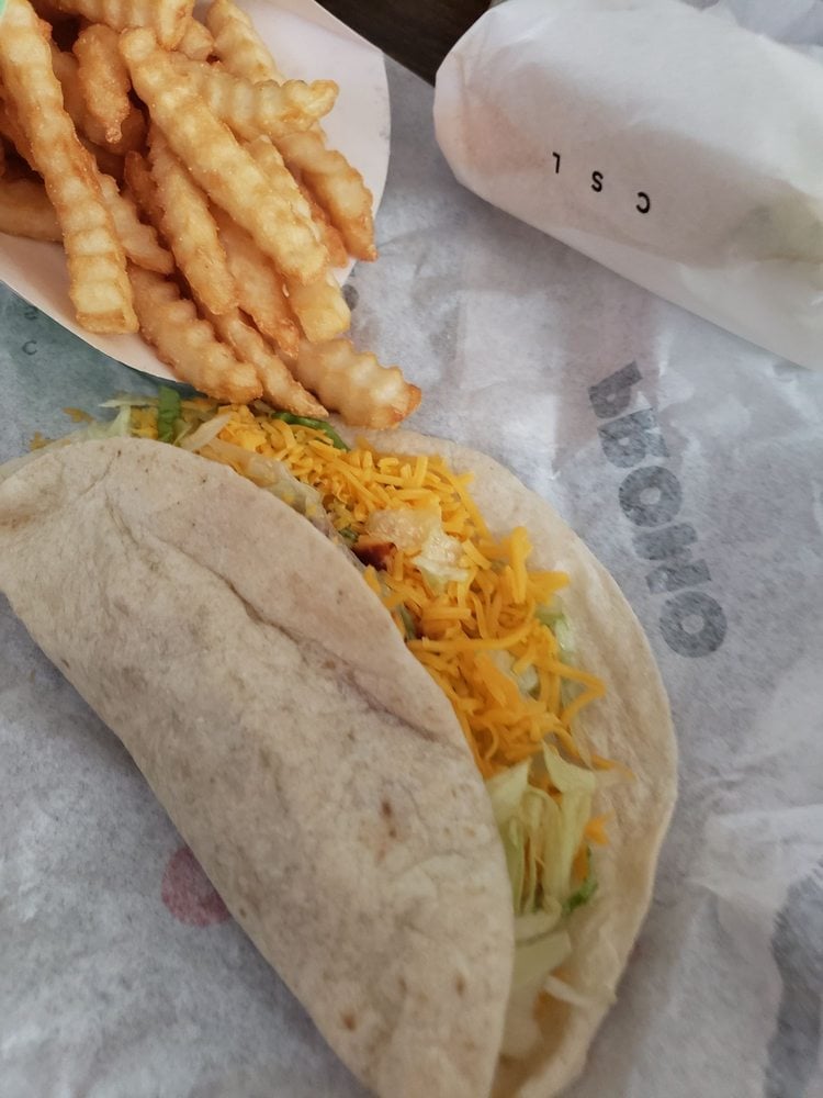 A soft taco filled with shredded lettuce and cheddar cheese sits on a wrapper next to crinkle-cut French fries and a wrapped item in the background.