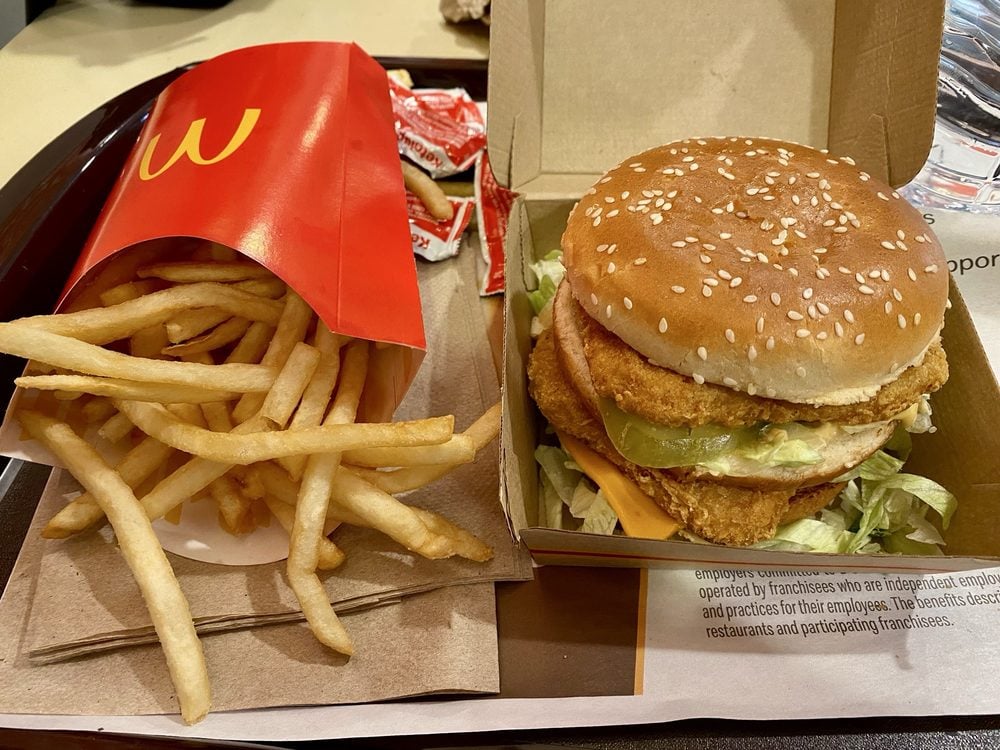 A McDonald’s meal on a tray with a box of French fries, a breaded fish sandwich with lettuce, cheese, and tartar sauce in an open container, ketchup packets, and a cup in the background.