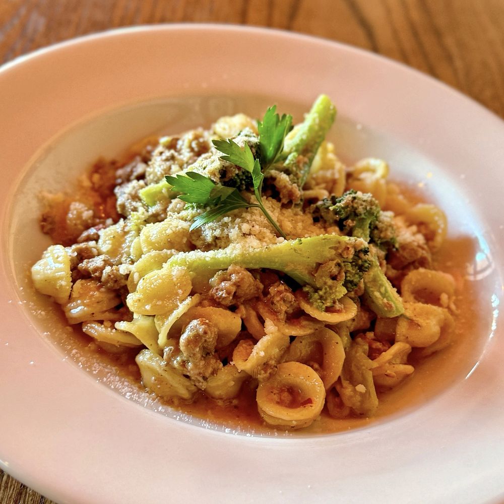 A white plate of orecchiette pasta with crumbled sausage, broccoli, grated cheese, and fresh parsley—just like the dishes served at the Best Italian Restaurants—sits on a wooden table.