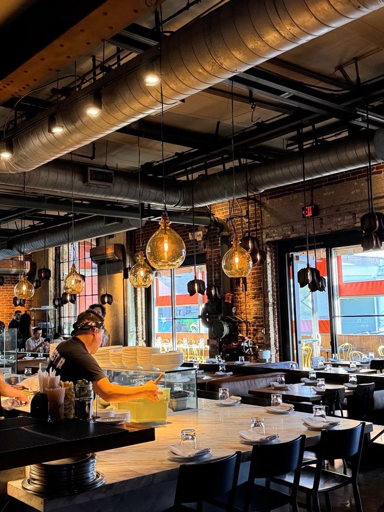A warmly lit, modern restaurant with exposed brick walls, large windows, and industrial ceiling pipes—perfect for those seeking the best Italian restaurants. A staff member works behind the counter; tables are set with glasses and napkins for guests.