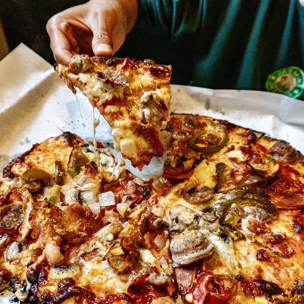 At one of the Best Italian Restaurants, a person lifts a cheesy slice of loaded pizza topped with sausage, onions, jalapeños, and other delicious toppings from a whole pizza on parchment paper.