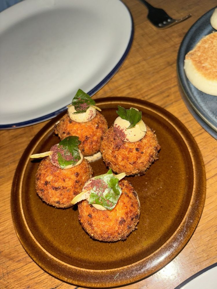 Four round, golden-brown, breaded appetizers are arranged on a brown plate, each topped with creamy sauce and fresh herbs—a tempting starter often found at the Best Italian Restaurants. An empty plate and fork are in the background.