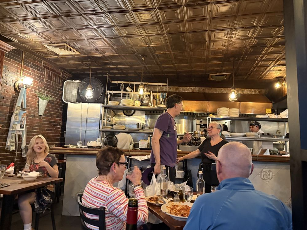 A lively restaurant scene with people dining and chatting, reminiscent of the Best Italian Restaurants. An open kitchen bustles in the background, while decorative ceilings and walls lined with shelves and plants add charm.