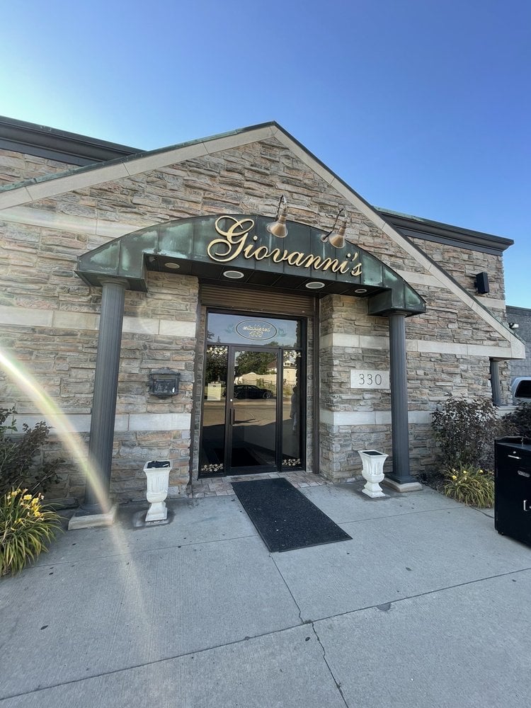Exterior of Giovanni’s, one of the Best Italian Restaurants, features a stone facade, green awning, and gold script sign above the entrance. Two white planters and a black mat frame the door, with the address number 330 clearly visible.