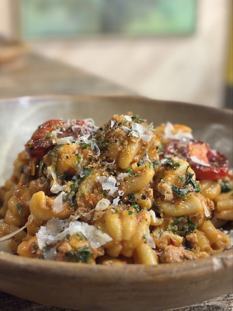 A close-up of a bowl of pasta with a creamy sauce, topped with grated cheese, greens, and roasted tomatoes, served in a rustic brown dish—just like you’d find at the Best Italian Restaurants.
