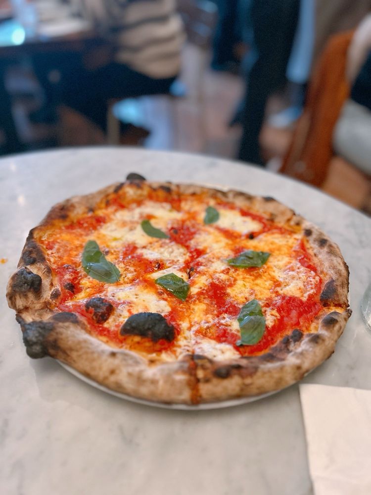 A margherita pizza with tomato sauce, melted cheese, and fresh basil leaves sits on a round plate on a marble table—a classic dish often found at the Best Italian Restaurants. The crust is browned and slightly charred, with diners and chairs blurred in the background.