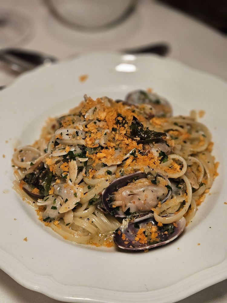 A plate of spaghetti with clams, garnished with chopped herbs and golden breadcrumbs, served in a white bowl on a set table—just like you'd find at the best Italian restaurants.