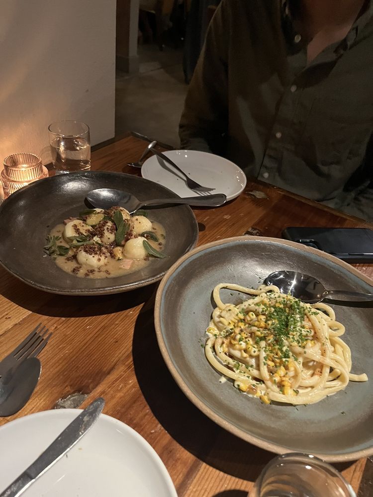 Two plates of pasta dishes sit on a wooden table at one of the Best Italian Restaurants, with utensils, empty plates, a glass of water, lit candle, and a person in a green shirt partially visible at the table.
