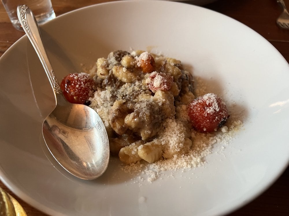 A white bowl of gnocchi topped with grated cheese, three roasted cherry tomatoes, and creamy sauce, served with a metal spoon—just what you'd expect at the best Italian restaurants.