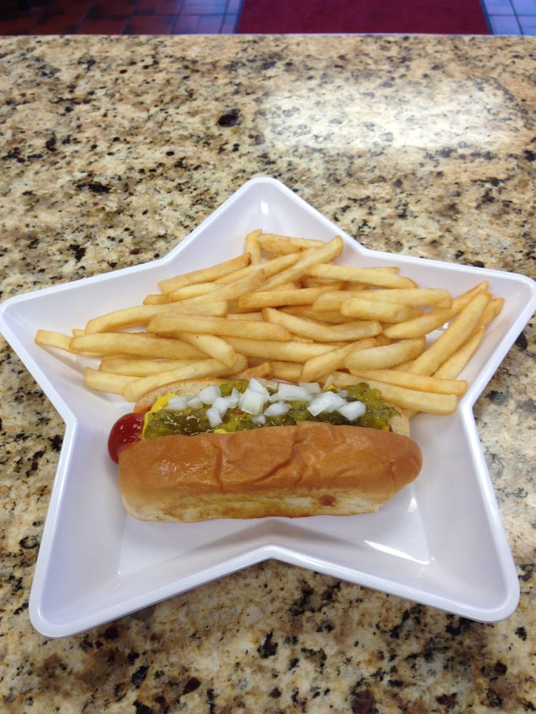 A star-shaped white plate holds a Fast-Food Hot Dog topped with onions, relish, mustard, and ketchup, alongside a serving of French fries, on a speckled countertop.