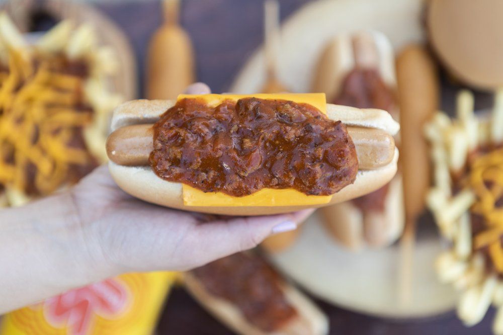 A hand holds a Fast-Food Hot Dog in a bun topped with cheddar cheese and chili. In the background, corndogs, fries with shredded cheese, and another hot dog sit on a wooden surface.