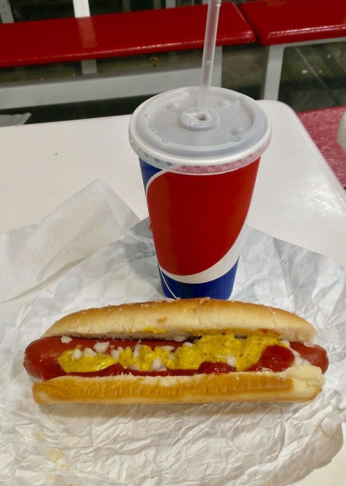A Fast-Food Hot Dog topped with ketchup, mustard, and chopped onions sits on foil next to a large soft drink in a red, white, and blue cup with a straw, on a white table with red bench seating in the background.