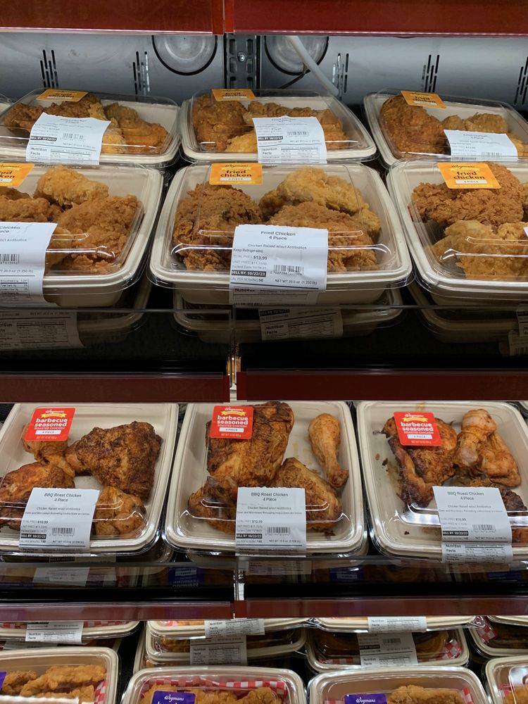 Plastic containers of fried, baked, and rotisserie chicken pieces are stacked on grocery store shelves, each package labeled with white or red stickers indicating product details and prices.