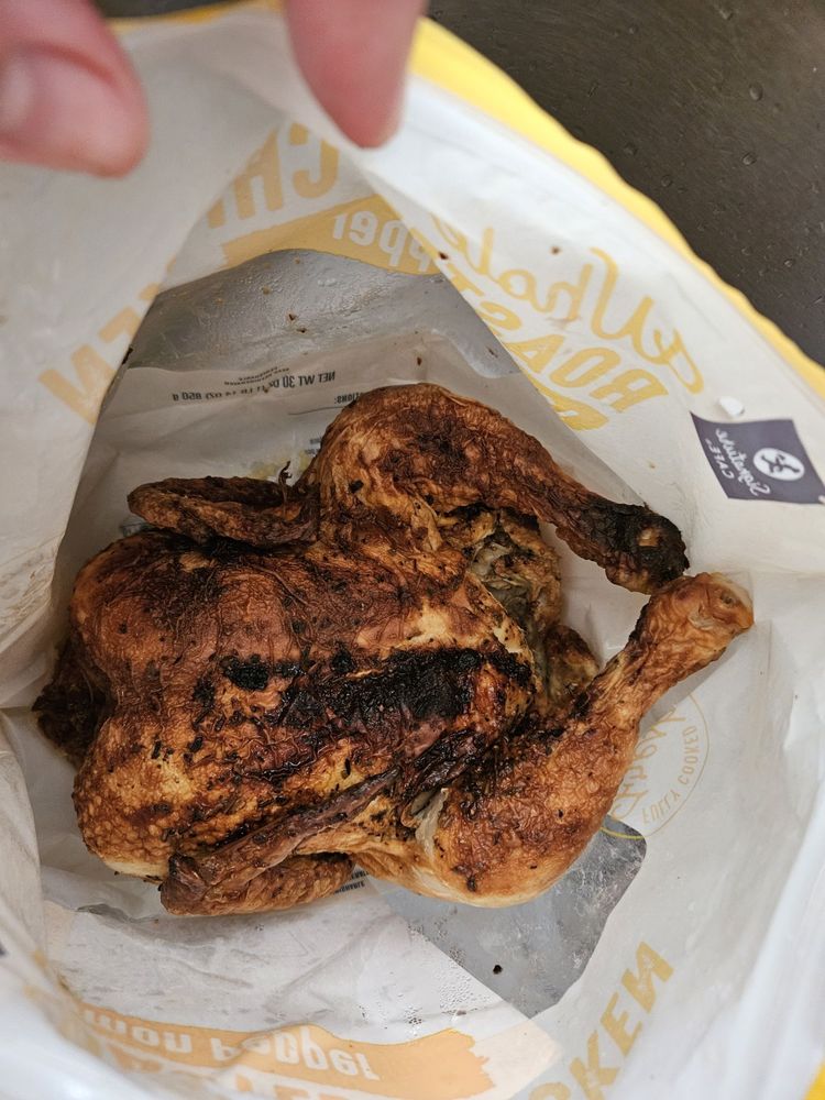 A delicious Rotisserie Chicken with browned skin rests inside a partially opened white and yellow paper bag, viewed from above. A hand is holding the edge of the bag open.