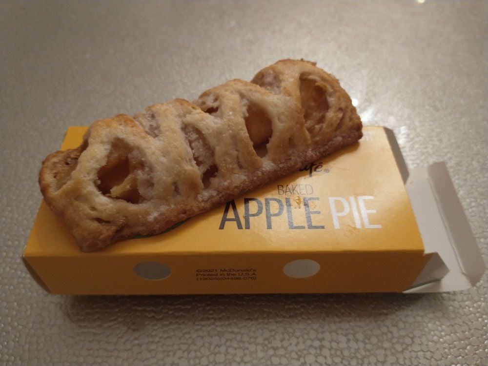 A McDonald's baked apple pie sits on top of its yellow packaging, showing its golden-brown crust with visible apple filling. The package is labeled "Baked Apple Pie" on a textured surface.