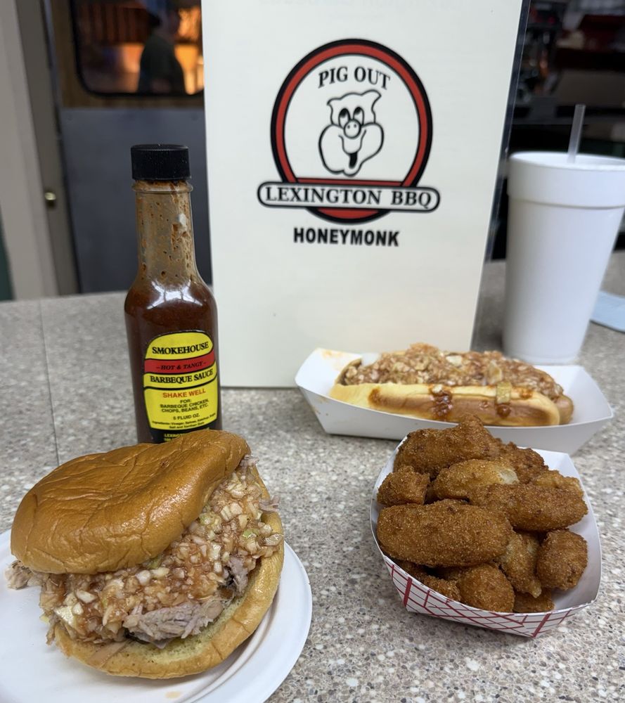 A tray with a chopped barbecue sandwich, fried hush puppies, a chili dog, a bottle of barbecue sauce, and a drink cup sits on a counter in front of a Lexington BBQ "Pig Out" menu—perfect for classic road trip food stops.