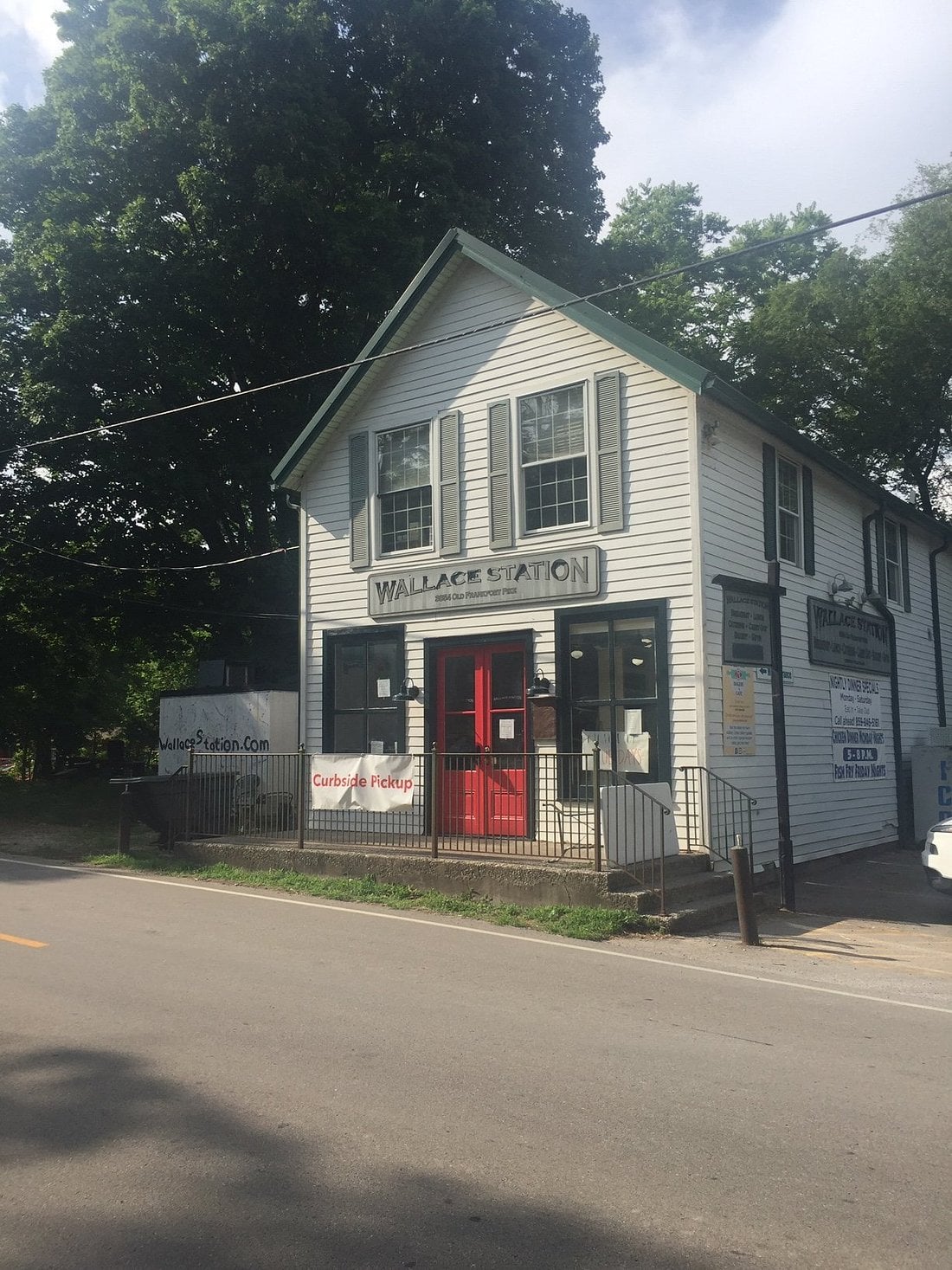 A two-story white building with green trim labeled "Wallace Station" sits beside a quiet road—an ideal spot for road trip food stops—with red double doors, curbside pickup signs, and trees under a partly cloudy sky in the background.