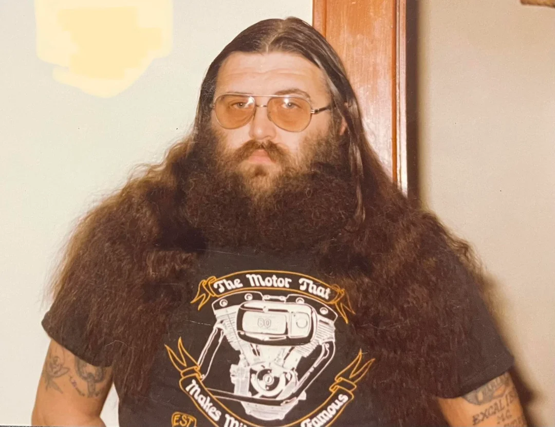 A man with long brown hair, a thick beard, and glasses stands indoors. He wears a black T-shirt with a graphic of a motorcycle engine and text. His arms have tattoos, and he looks directly at the camera.