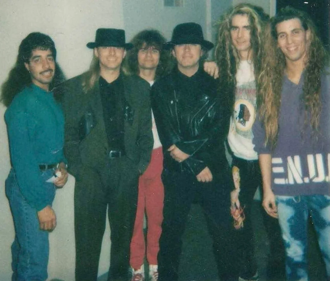 Six men stand together indoors, posing for a photo. Most have long hair; two wear black hats and jackets. The others are dressed in casual clothes, including jeans, a graphic sweater, and a suit. All are smiling or looking at the camera.