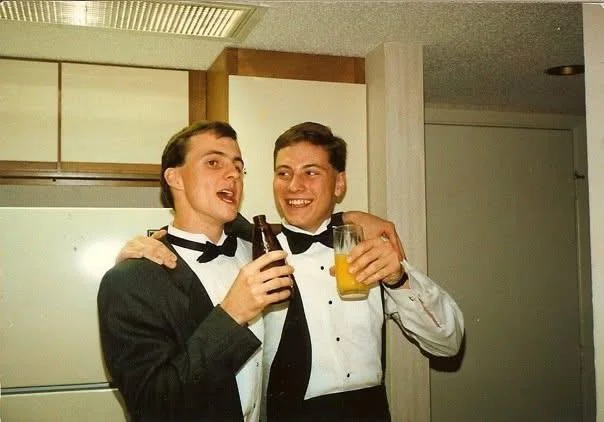 Two young men in tuxedos smile and pose together indoors, each holding a drink. One has his arm around the other, and they appear to be celebrating or enjoying a party.