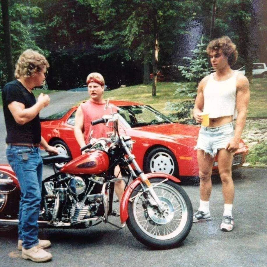 Three people stand by a red motorcycle and a red sports car outdoors. One person wears a crop top and cutoff shorts, another wears a red tank top, and the third wears jeans and a black shirt. Trees are in the background.
