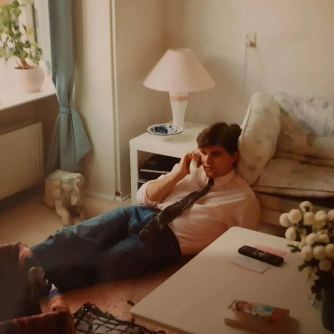 A man in a white shirt and tie sits on the floor, leaning against a sofa, talking on a phone. A coffee table with a book, remote, and flowers is in front of him. The room is softly lit with a table lamp and a window.
