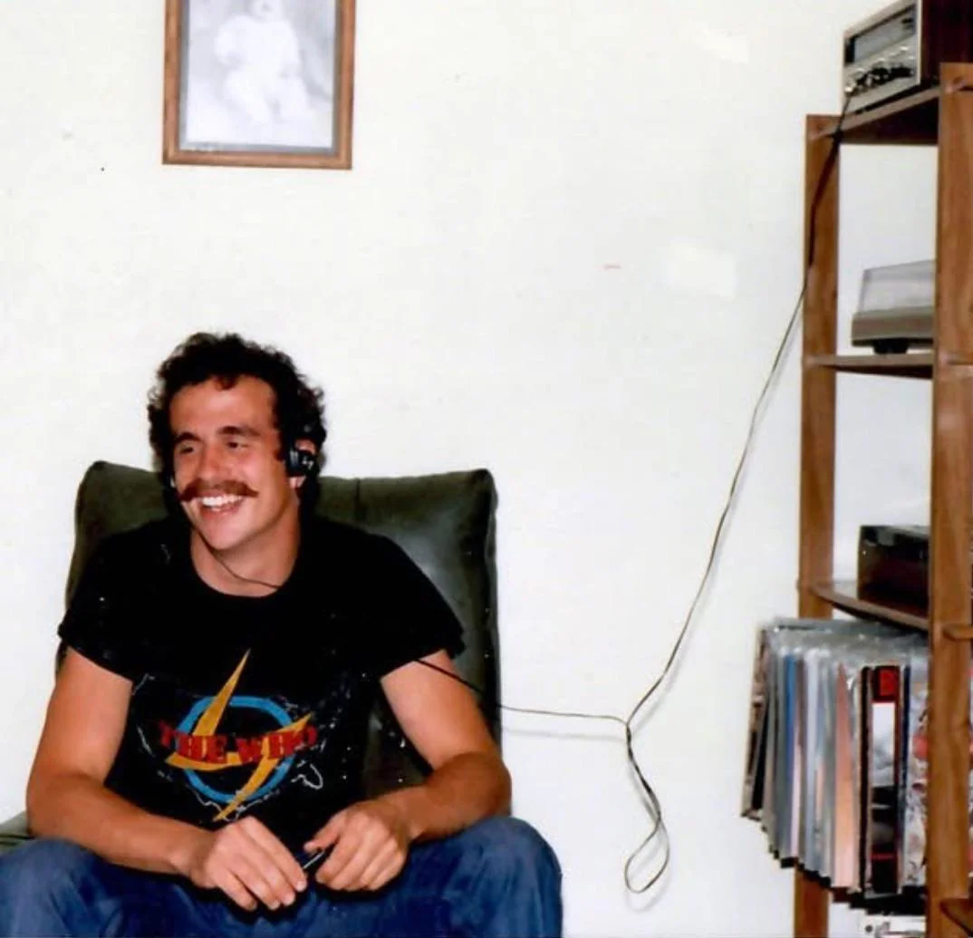 A smiling man with a mustache sits in a chair, wearing a headset and a black t-shirt with a colorful graphic. Beside him is a wooden shelf holding vinyl records, a stereo, and other audio equipment.