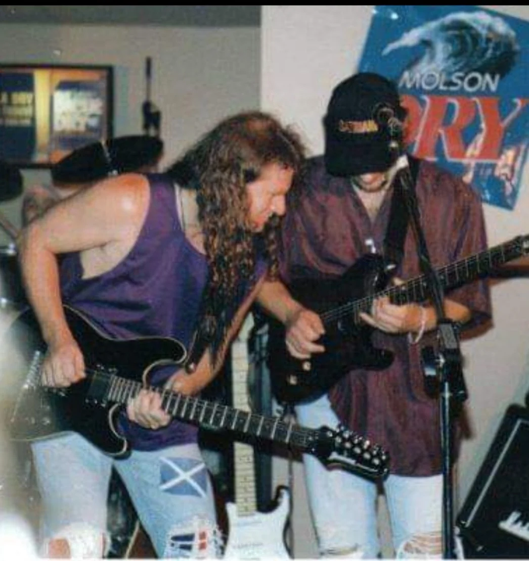 Two musicians with long hair play electric guitars together on stage. Both wear casual clothes, including ripped jeans, while intently focusing on their instruments. A Molson Dry beer poster is visible in the background.