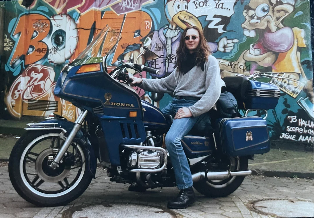 A person with long hair, sunglasses, jeans, and a gray sweater sits on a blue Honda motorcycle parked in front of a colorful graffiti-covered wall.