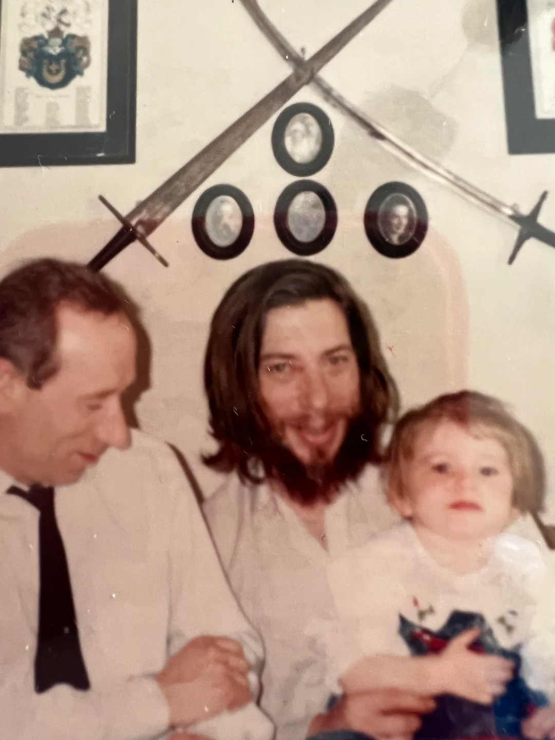 Three people sit close together indoors. An older man in a white shirt and tie looks down, a bearded man with long hair smiles, and a young child sits in front. Two swords and framed portraits hang on the wall behind them.