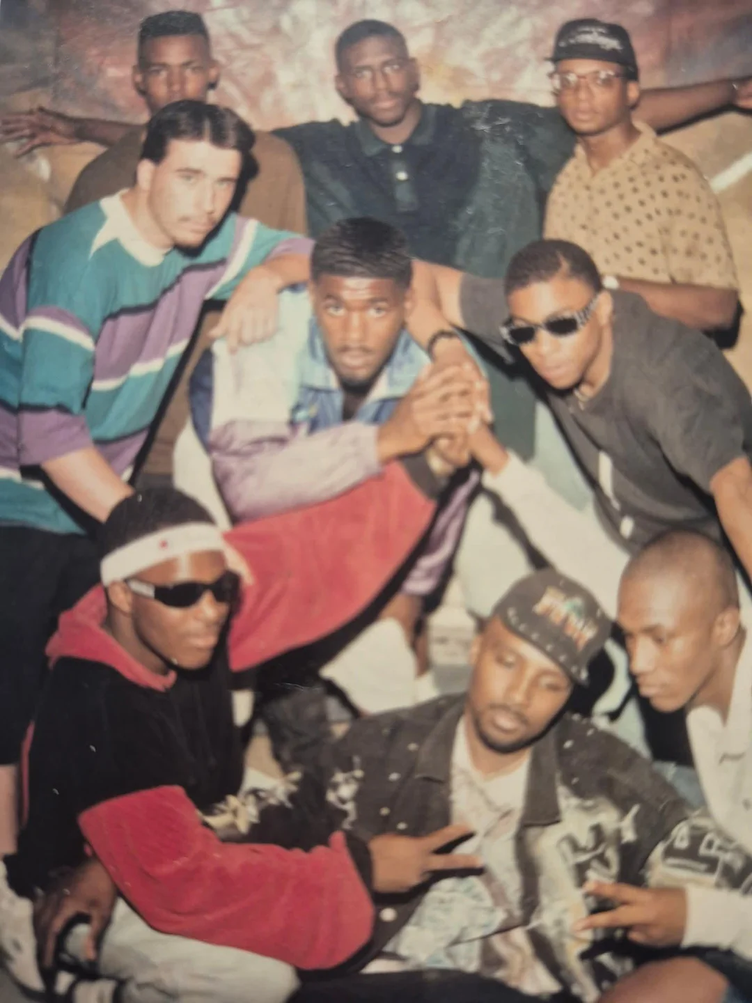 Nine young men posing together, some crouched in front and others standing in back. Most are wearing sunglasses, casual streetwear, and caps, showing relaxed and confident expressions.