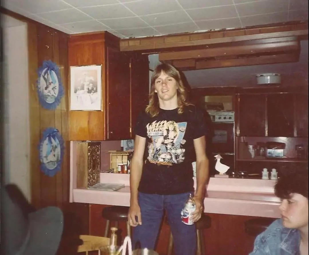 A young man with long hair stands in a wood-paneled kitchen, wearing a black graphic T-shirt and jeans, holding a drink. Another person sits nearby, partly visible. Retro decor and pink countertops are in the background.