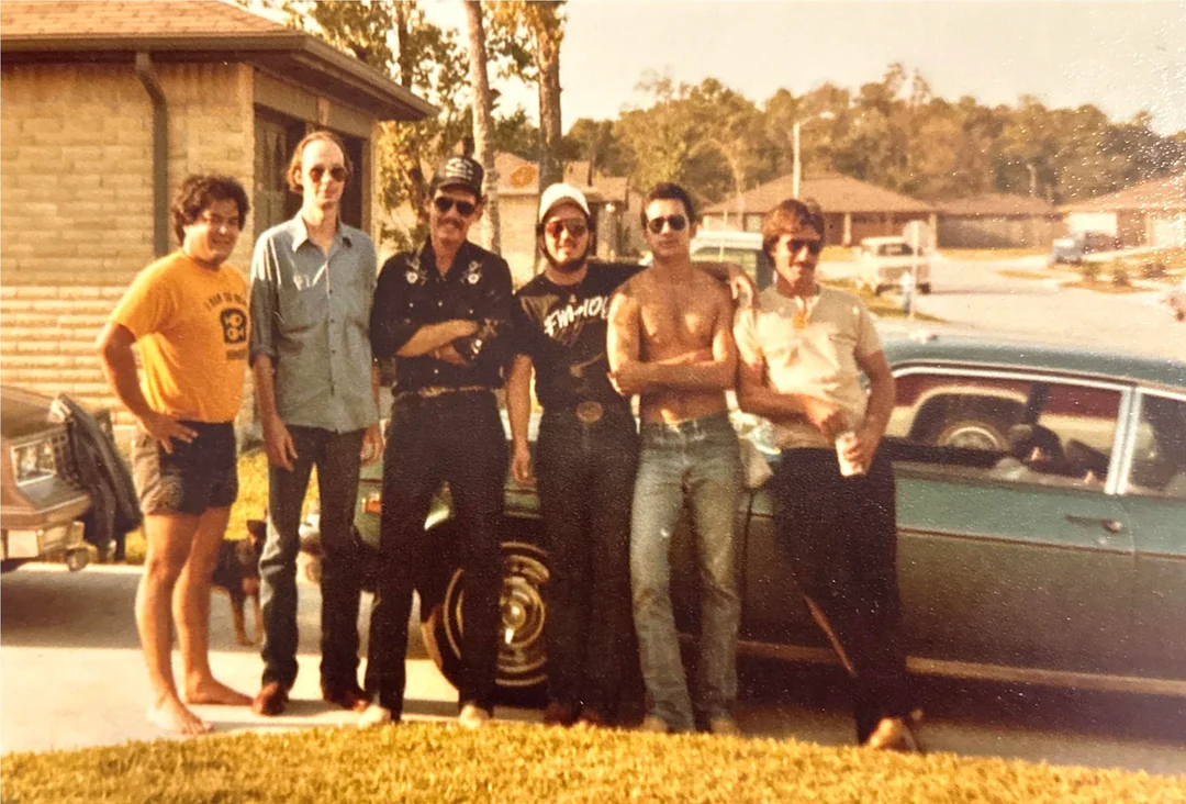 Six men pose casually in front of a vintage car parked in a suburban neighborhood. Some wear sunglasses; one is shirtless, and several wear hats. The image has a warm, nostalgic, vintage feel. 80s and 90s dads. 