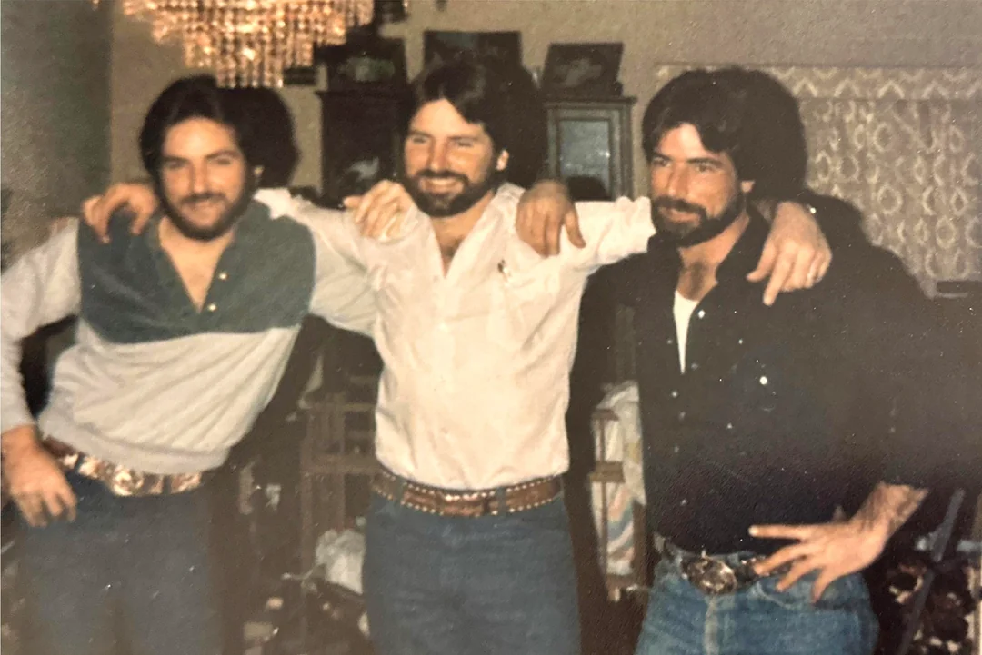 Three men with beards and long dark hair stand close together indoors, smiling at the camera. They wear casual button-down shirts, jeans, and large belt buckles, with arms around each other's shoulders.