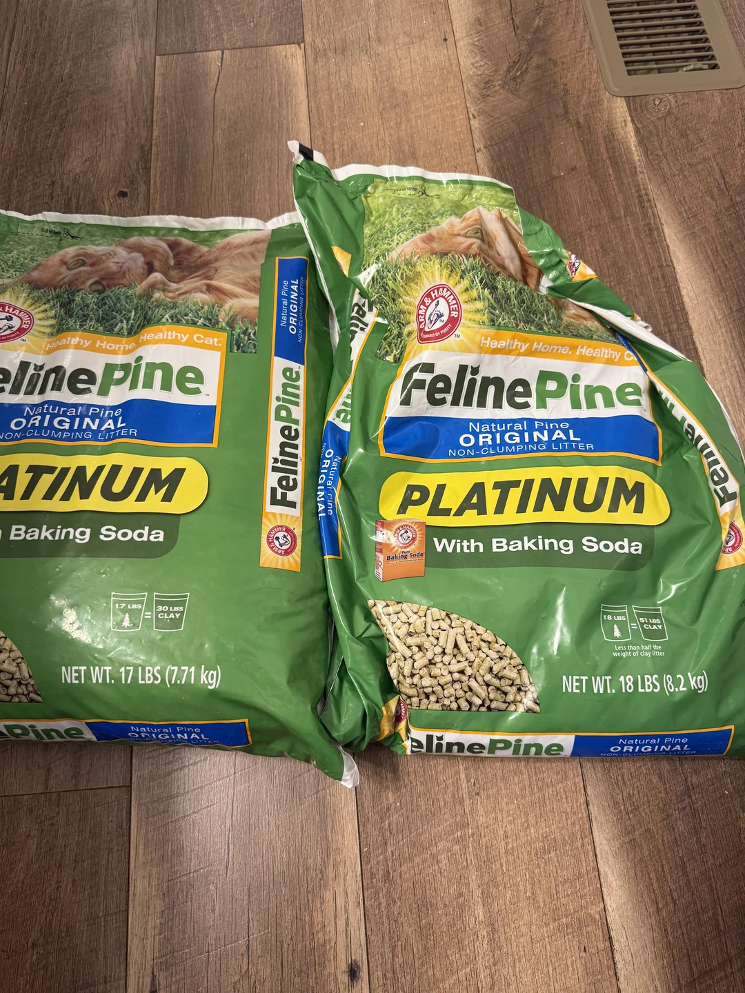 Two green bags of Feline Pine Platinum cat litter with baking soda sit on a wooden floor. One bag weighs 17 lbs and the other 18 lbs. Both show images of pine and cats, and feature yellow and blue branding.