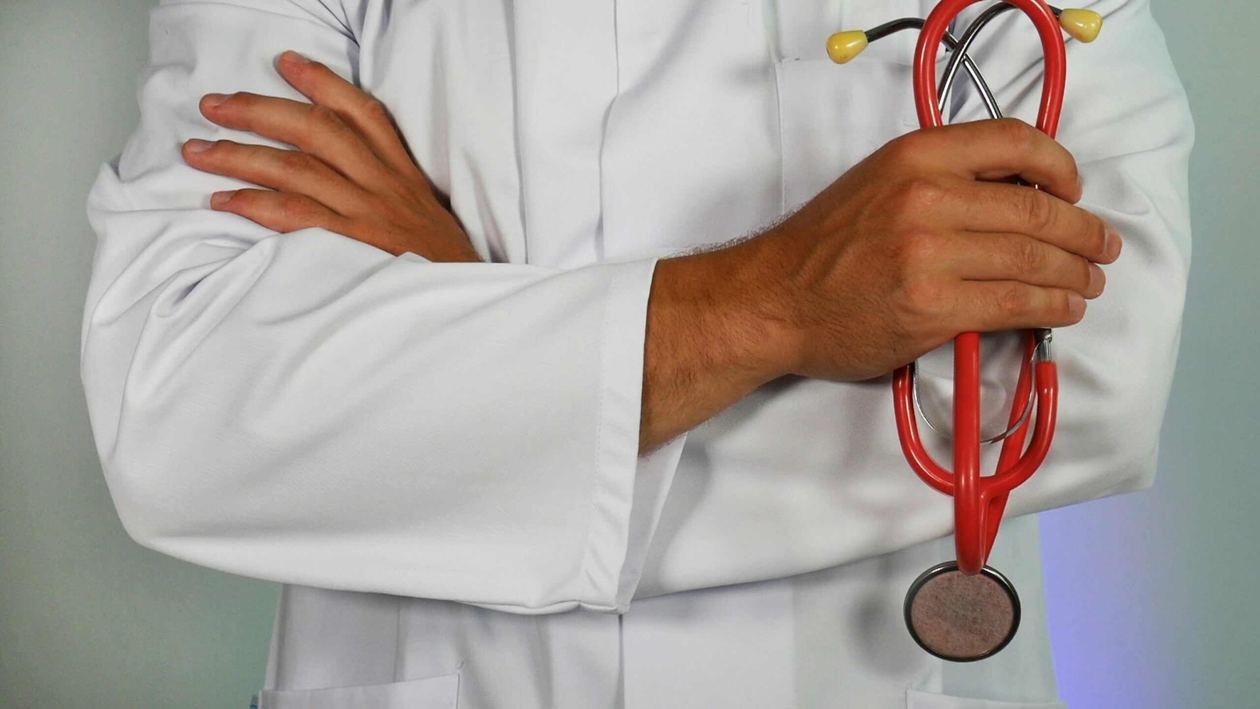A person in a white lab coat stands with arms crossed, holding a red stethoscope in one hand, suggesting a healthcare professional. The face is not visible.