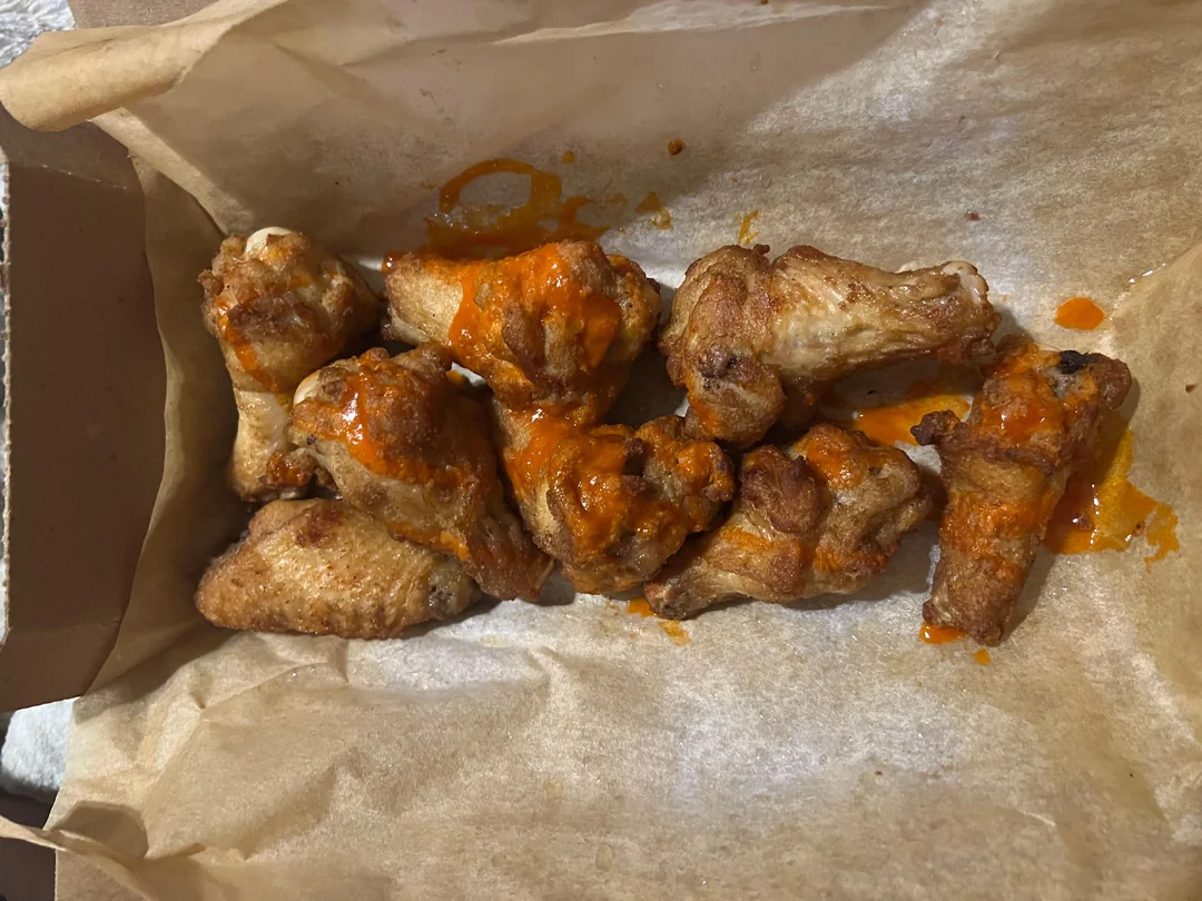Eight cooked chicken wings with a light coating of orange buffalo sauce are placed on brown parchment paper inside a cardboard takeout container.