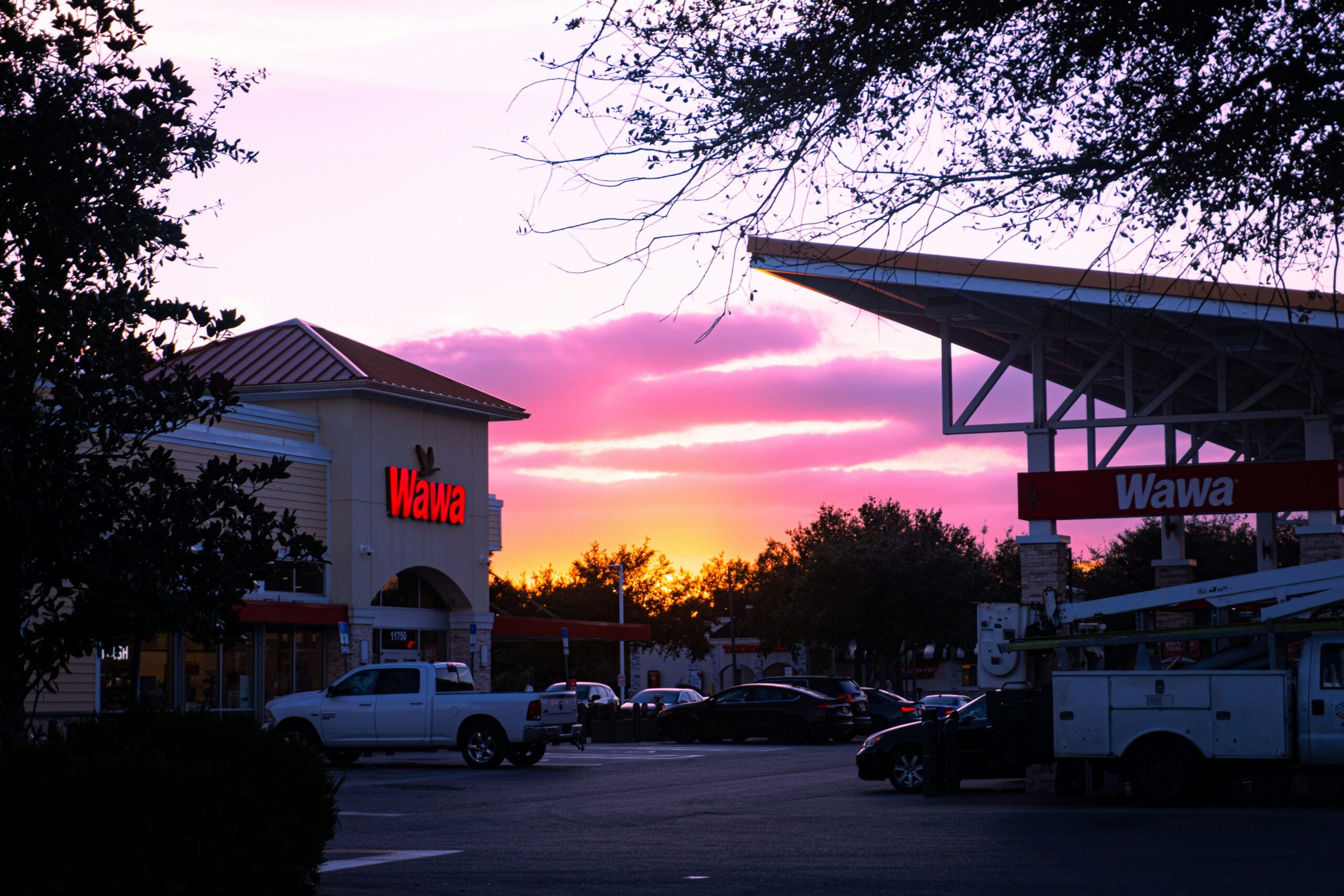 A Wawa gas station and convenience store at sunset, with a colorful sky of pink, purple, and orange hues. Cars and a utility truck are parked in the lot, and trees frame the scene.
