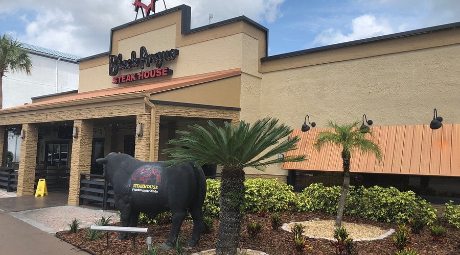 A beige building houses the Black Angus Steakhouse, with a black bull statue and palm plants in front, and a red bull statue on the roof under a cloudy sky.