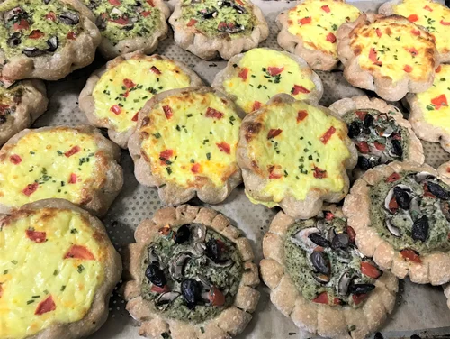 A tray filled with small, round savory tarts topped with a variety of ingredients including cheese, chopped red peppers, black olives, mushrooms, and herbs. The tarts have a scalloped crust.