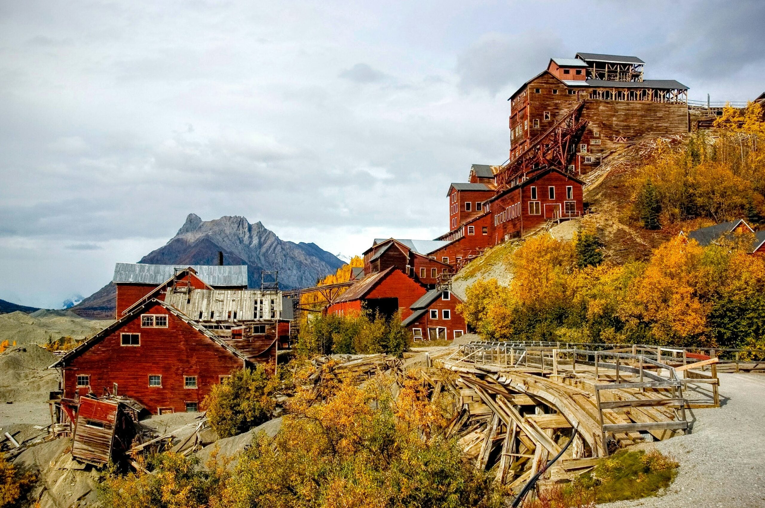 Abandoned red wooden mining buildings sit on a hillside surrounded by autumn trees, with rugged mountains and a cloudy sky in the background. Wooden walkways and old structures are visible in the foreground.