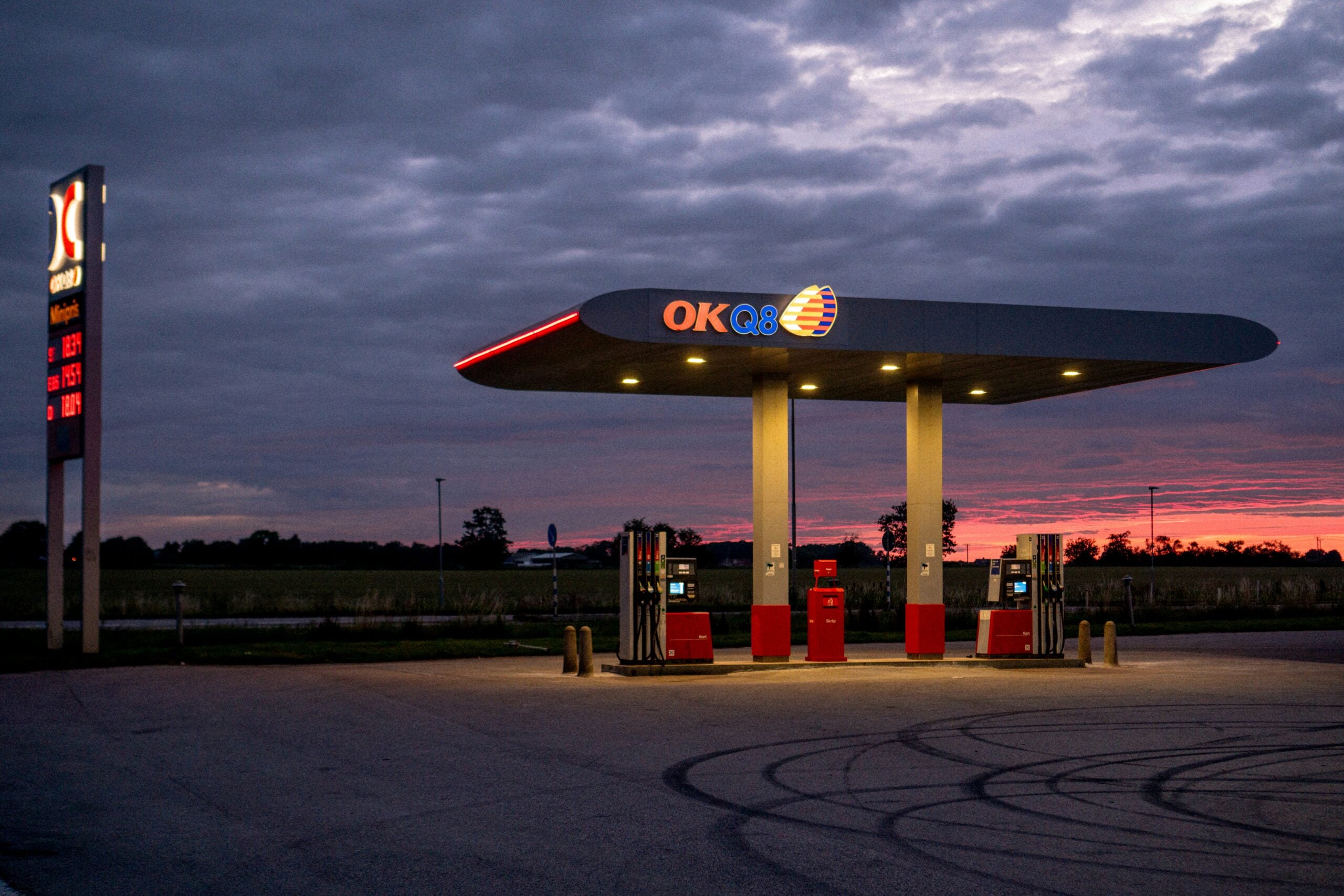 An empty OKQ8 gas station is lit up at dusk, with a cloudy sky and a red-orange sunset in the background. The price sign glows on the left, and fields stretch out beyond the station.