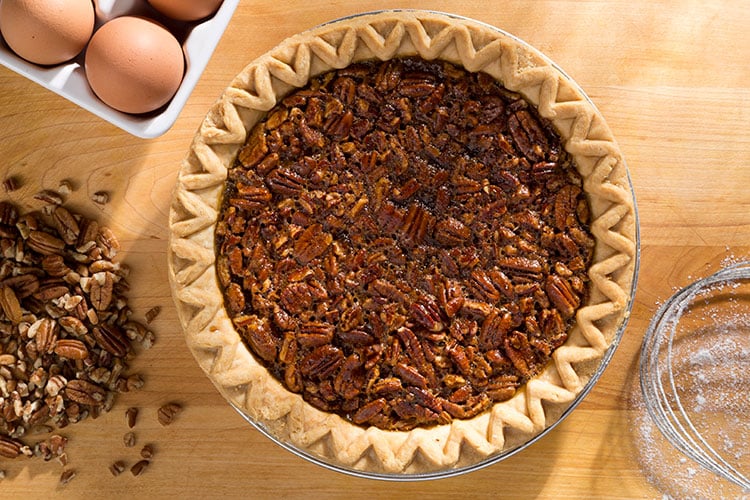 A pecan pie with a golden, crimped crust sits on a wooden surface, surrounded by a bowl of eggs, a pile of pecans, and a glass bowl with some flour.
