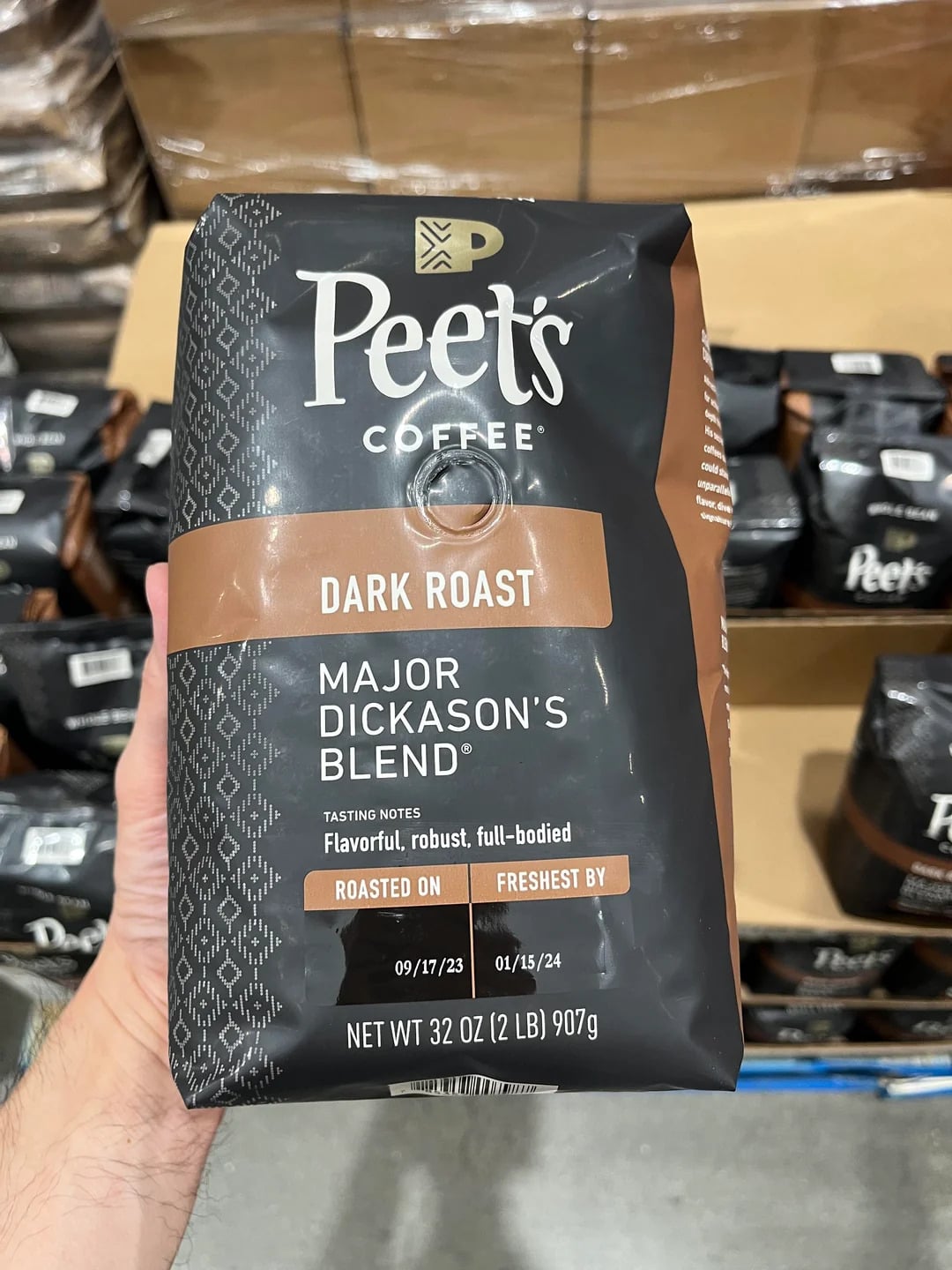 A hand holds a 32 oz bag of Peet's Coffee, Dark Roast, Major Dickason's Blend—one of the top coffee brands—in a store. The black and brown packaging features roast and freshness dates. Other coffee bags are visible in the background.