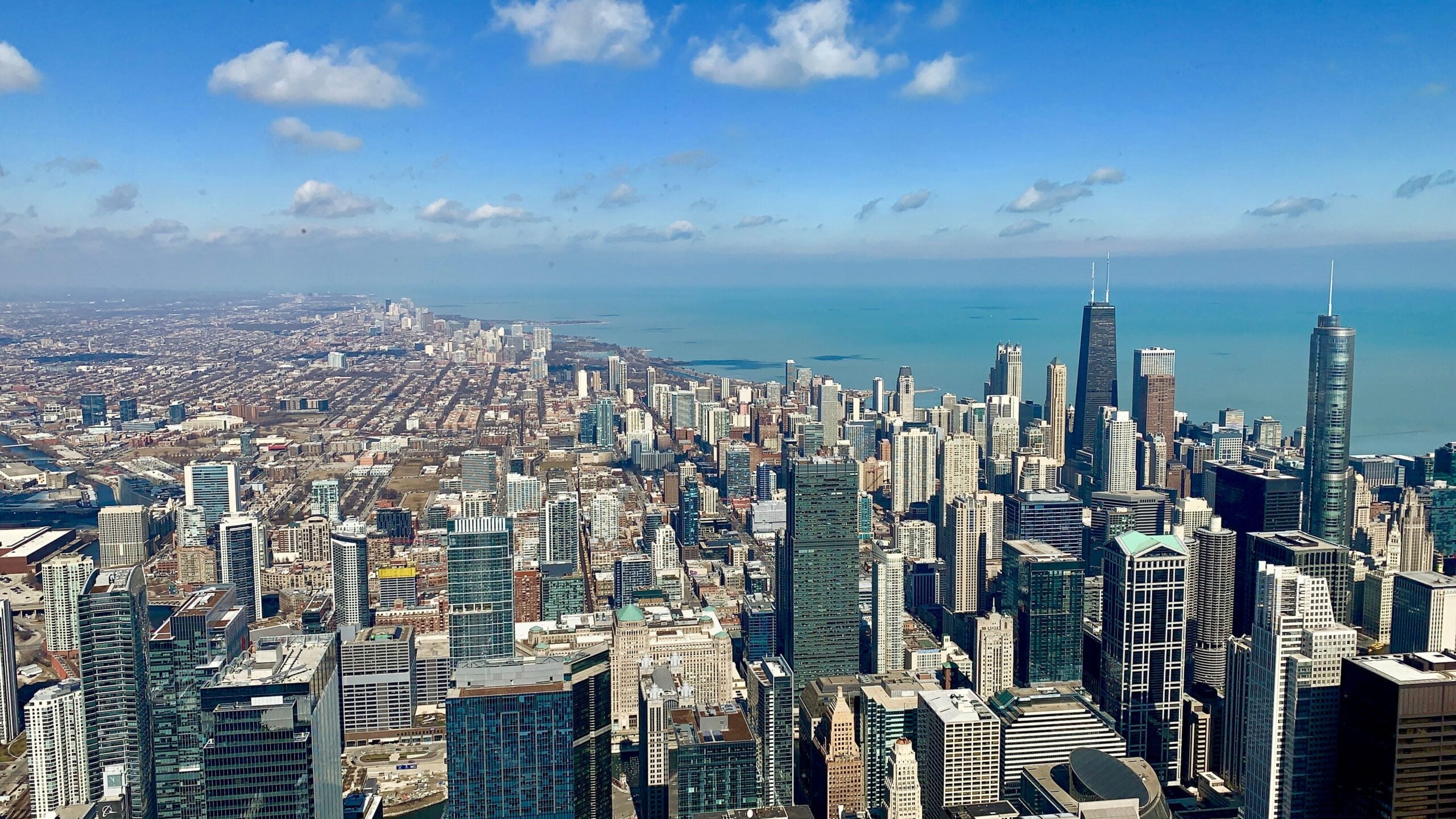 Aerial view of downtown Chicago with tall skyscrapers, Lake Michigan in the background, and a partly cloudy blue sky above the city highlights an urban landscape where Cost of Living Relief is increasingly essential.