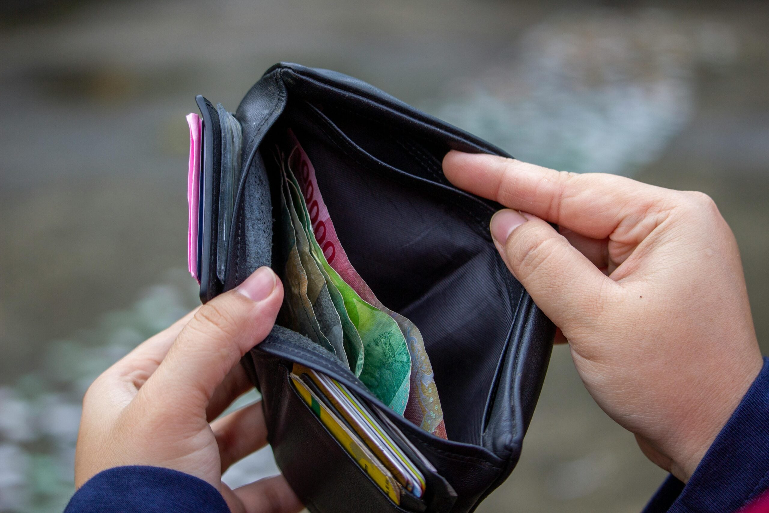A person holding an open wallet, showing a few folded paper bills and several cards inside, with a blurred outdoor background.