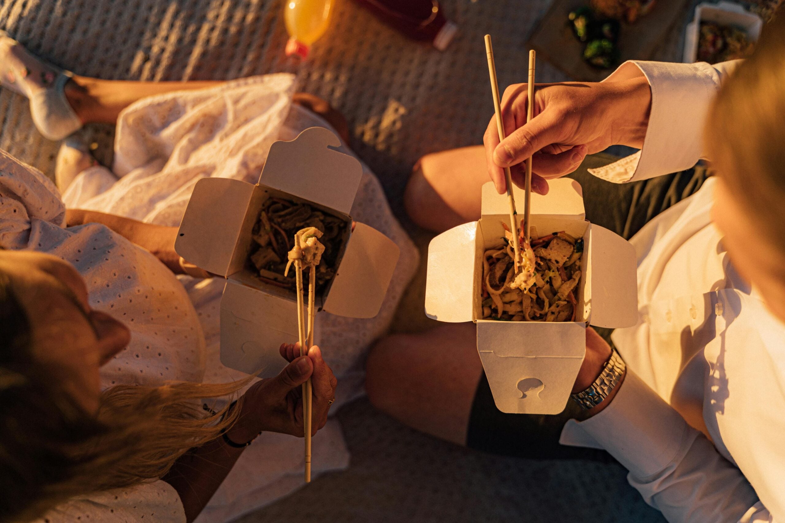 Two people sitting on a blanket outdoors, eating noodles from takeout boxes with chopsticks. Sunlight creates a warm, relaxed atmosphere, and a drink and some food are visible in the background.