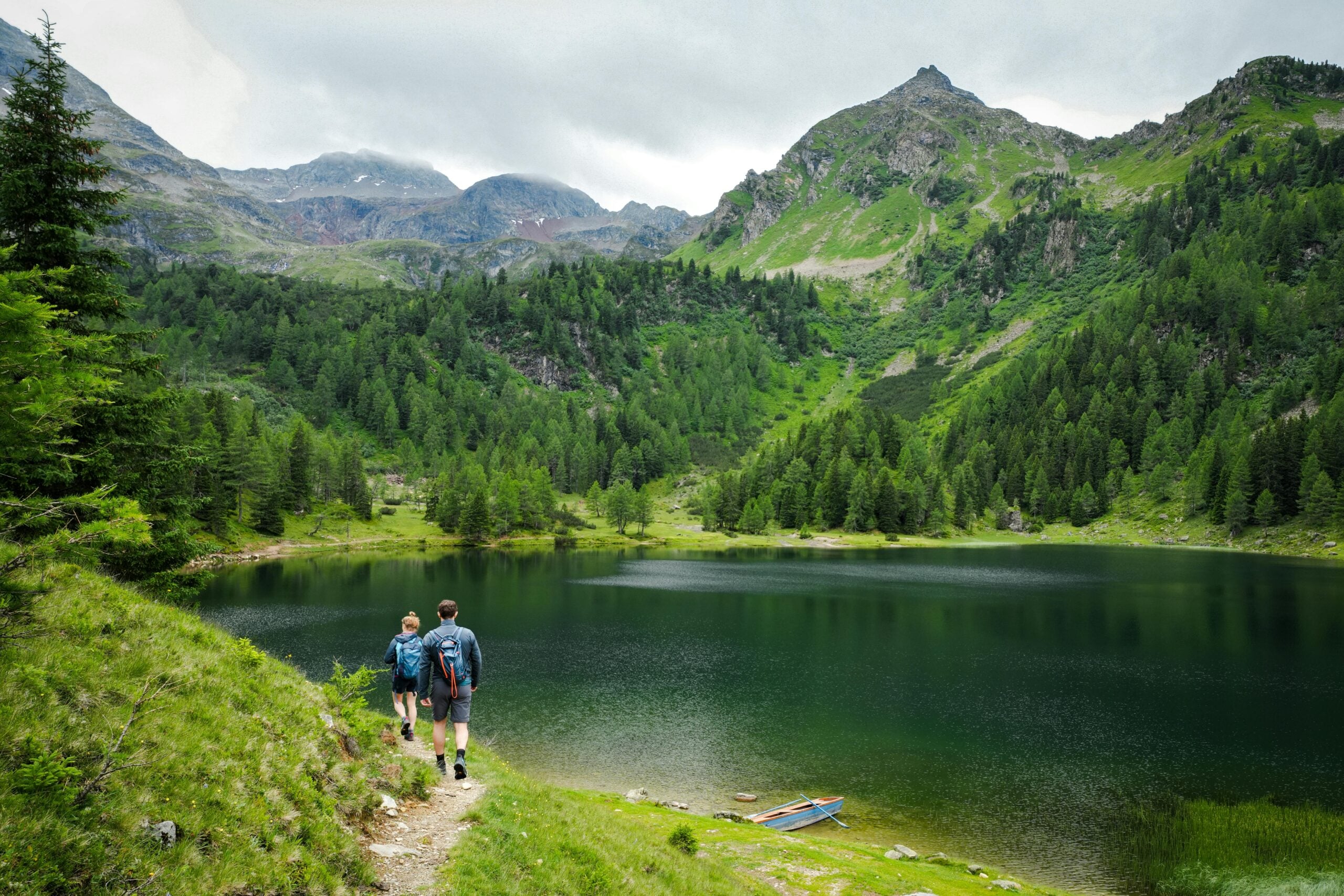 Two hikers walk along a grassy trail beside a calm mountain lake, surrounded by lush green trees and tall peaks under a cloudy sky. A small blue boat rests on the shore.